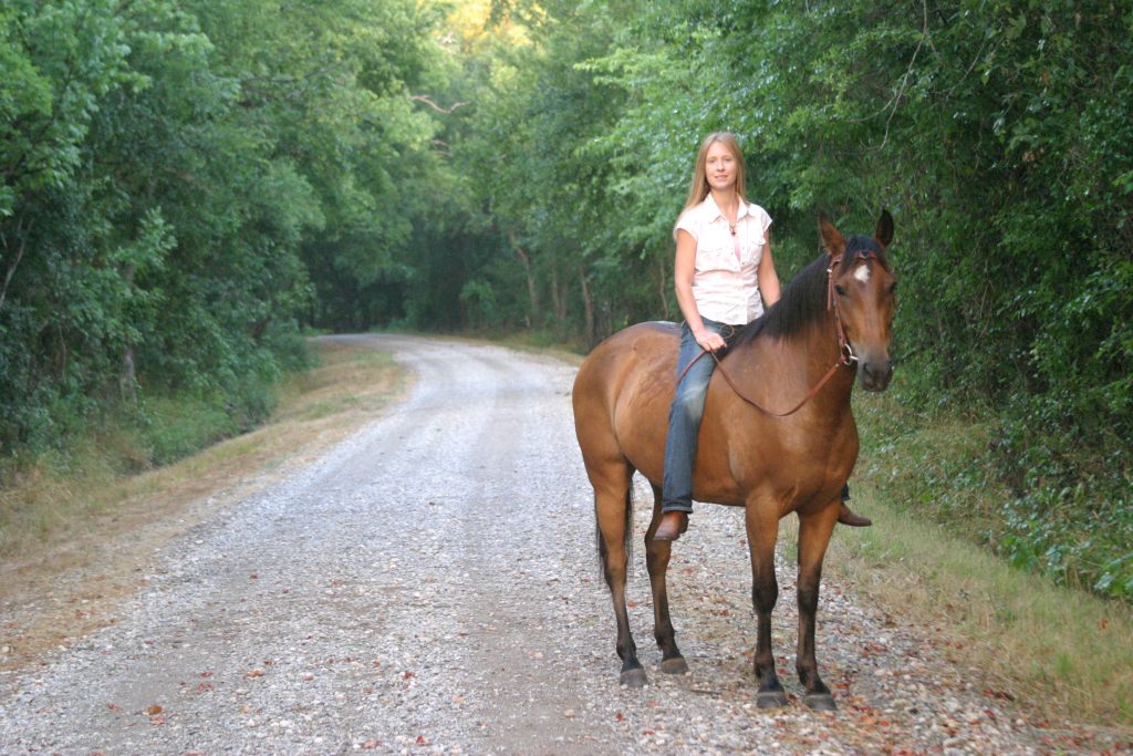 CharlieAnn riding a bay horse down a tree-lined gravel road